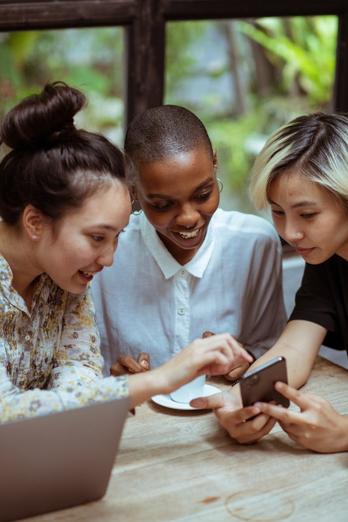 Three diverse women smile while using a smartphone in a cozy indoor setting.
